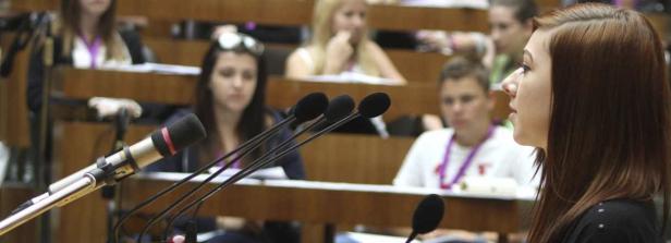 Eine junge Frau spricht vor einem Publikum in einem Hörsaal in ein Mikrofon.