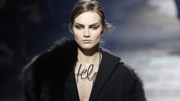 Ein Model mit dunklem Mantel und einer Halskette mit dem Wort „Help“.