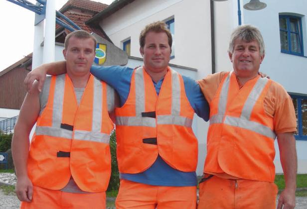 Drei Männer in orangefarbenen Warnwesten stehen nebeneinander.