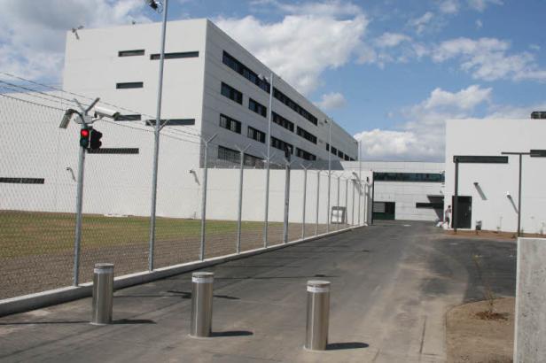 Ein modernes, weißes Gebäude mit einem Sicherheitszaun und versenkbaren Pollern davor.