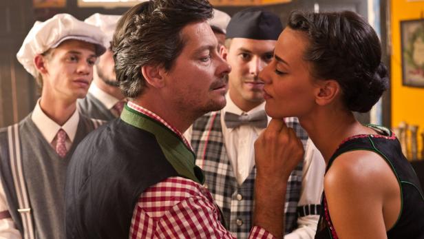 Eine Frau berührt das Kinn eines Mannes in Tracht, umgeben von weiteren Personen in traditioneller Kleidung.