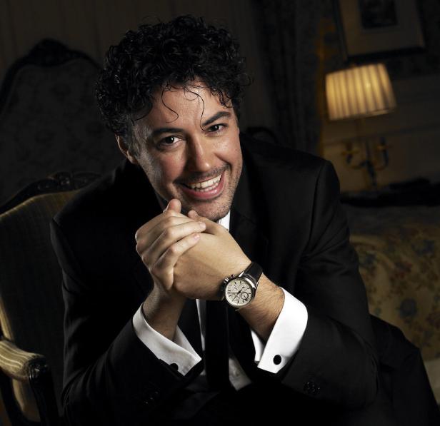 Ein lächelnder Mann im Anzug mit einer Armbanduhr sitzt in einem Sessel.