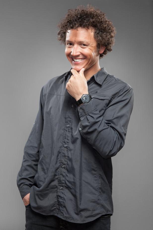 Ein Mann mit lockigem Haar lächelt und berührt sein Kinn.