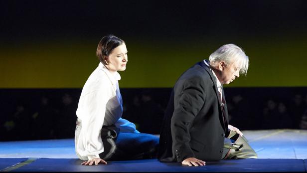 Eine Frau und ein Mann knien auf einer Bühne vor einem dunklen Hintergrund.