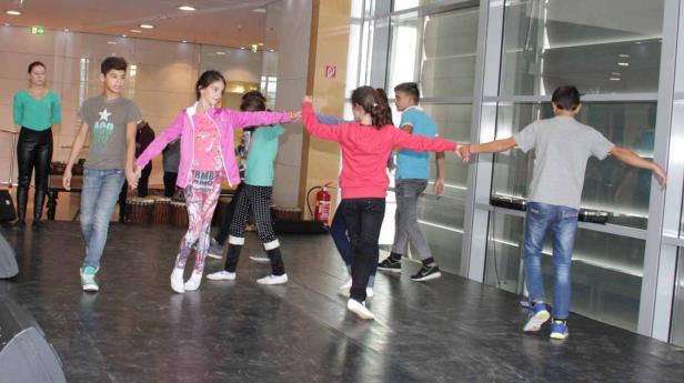 Eine Gruppe von Kindern tanzt auf einer Bühne, während eine Frau zusieht.