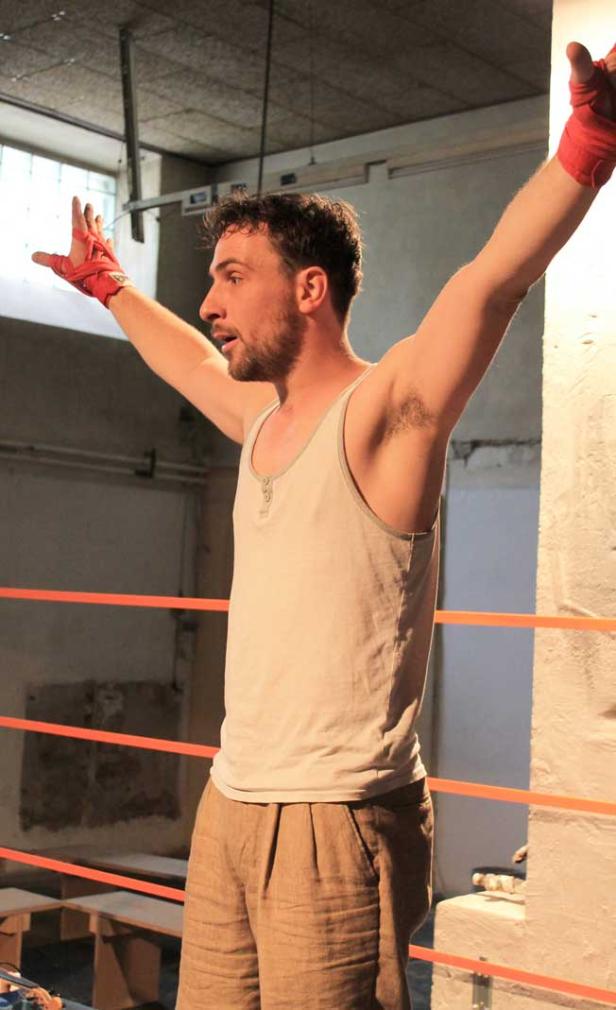 Ein Mann mit roten Bandagen an den Händen steht mit erhobenen Armen in einem Boxring.