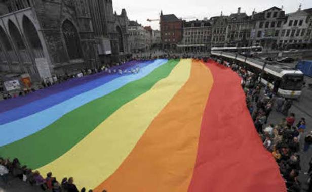 Eine riesige Regenbogenflagge wird in einer Stadt von Menschen gehalten.