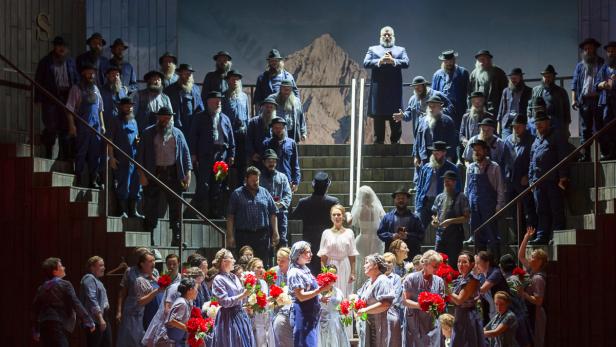 Eine Bühnenszene mit einer Gruppe von Männern und Frauen in traditioneller Kleidung, möglicherweise eine Opernaufführung.