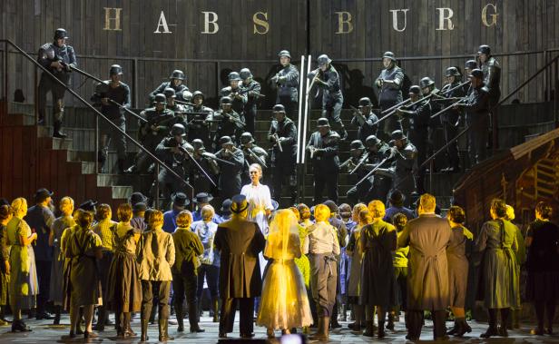 Eine Bühnenszene mit einer Menschenmenge vor einer Gruppe von Soldaten mit Gewehren unter dem Schriftzug „HABSBURG“.