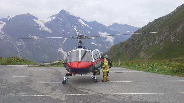 Ein rot-blauer Hubschrauber steht vor einer Bergkulisse auf einem Landeplatz.