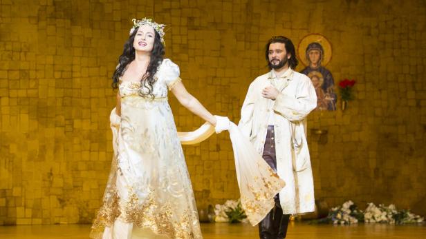 Eine Frau im weißen Kleid und ein Mann im Mantel stehen auf einer Bühne.