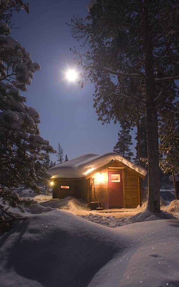 Eine schneebedeckte Hütte leuchtet im Mondschein zwischen Bäumen.