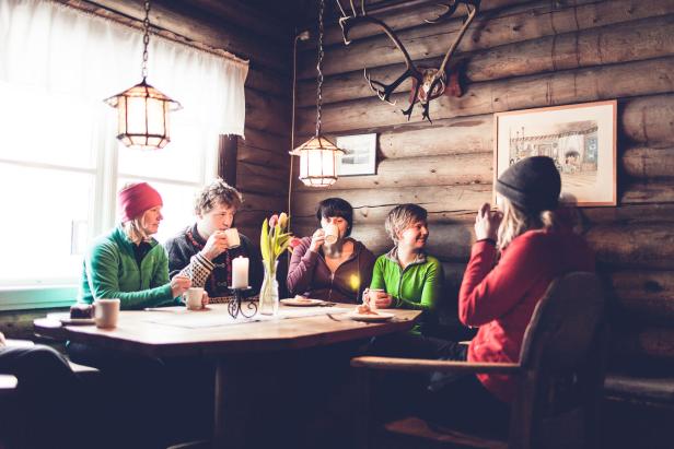 Fünf Personen sitzen in einer Blockhütte an einem Tisch und trinken Kaffee.