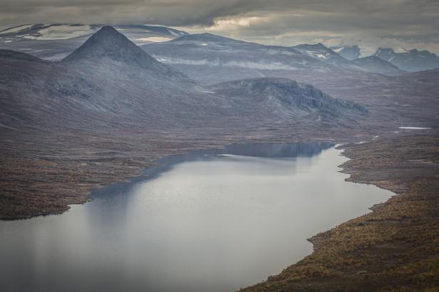 Ein stiller See liegt eingebettet in einer hügeligen Landschaft unter einem bewölkten Himmel.