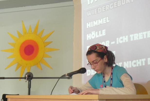 Eine junge Frau liest an einem Rednerpult vor, im Hintergrund eine Sonne und Text.