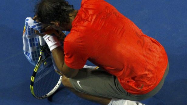 Ein Tennisspieler hockt erschöpft mit einem Handtuch im Gesicht auf dem Platz.