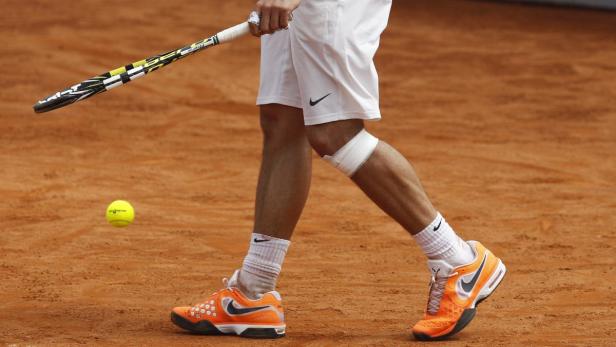 Ein Tennisspieler mit bandagiertem Knie auf einem Sandplatz mit Schläger und Ball.