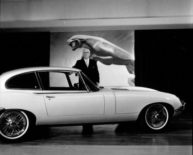 Ein Mann posiert neben einem Jaguar E-Type vor einem Jaguar-Logo.