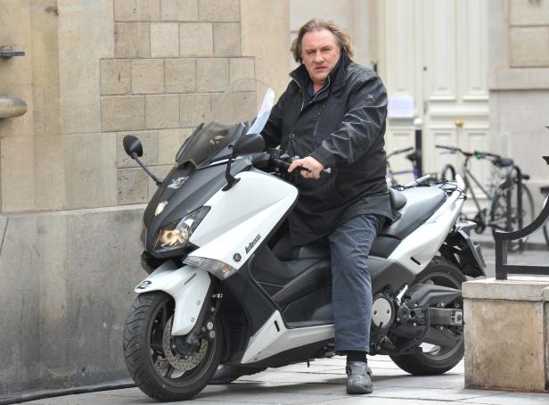 Gérard Depardieu sitzt auf einem Motorroller in einer städtischen Umgebung.