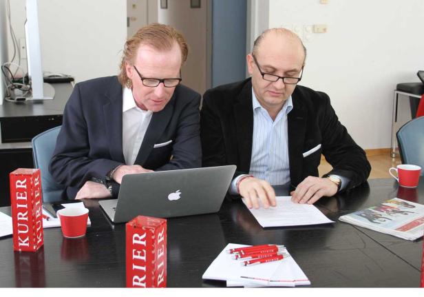 Zwei Männer sitzen an einem Tisch mit einem Laptop und Dokumenten, daneben Werbemittel des „Kurier“.