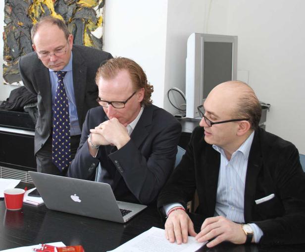 Drei Männer in Anzügen sitzen an einem Tisch und schauen auf einen Laptop.