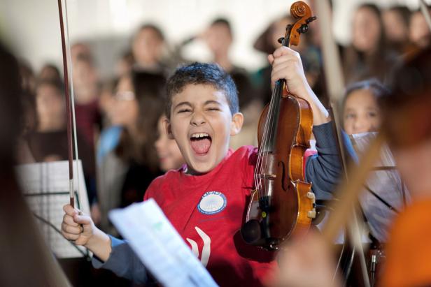 Ein aufgeregter Junge hält eine Geige in die Höhe, während er in einem Orchester spielt.