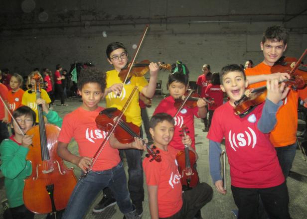 Eine Gruppe von Kindern mit Geigen und einem Cello posiert für ein Foto.
