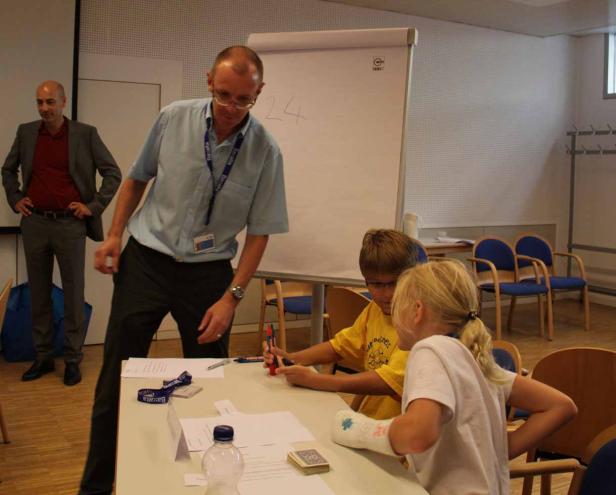 Ein Mann steht an einem Tisch, an dem zwei Kinder sitzen und arbeiten. Ein weiterer Mann steht im Hintergrund.