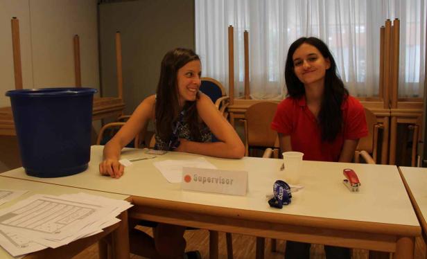 Zwei junge Frauen sitzen an einem Tisch mit einem Schild „Supervisor“.