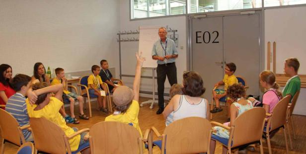 Ein Mann hält einen Vortrag vor einer Gruppe von Kindern und Erwachsenen in einem Raum.