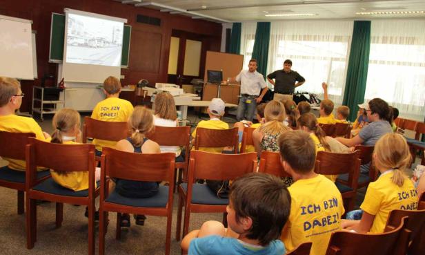 Zwei Männer präsentieren vor einer Gruppe von Kindern in einem Klassenzimmer.