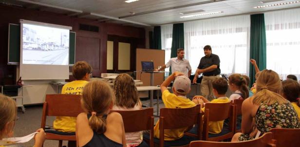 Zwei Männer präsentieren vor einer Gruppe von Kindern in einem Klassenzimmer.