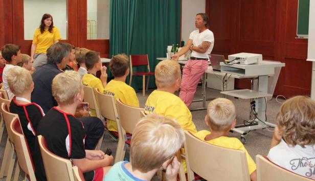 Ein Mann in rosa Hosen hält einen Vortrag vor einer Gruppe von Kindern.