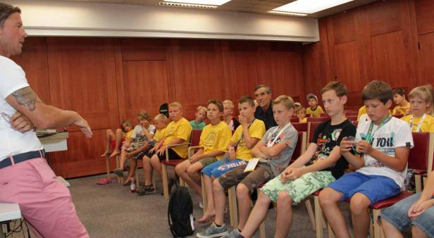 Ein Mann spricht vor einer Gruppe von Kindern in einem Raum mit Holzvertäfelung.