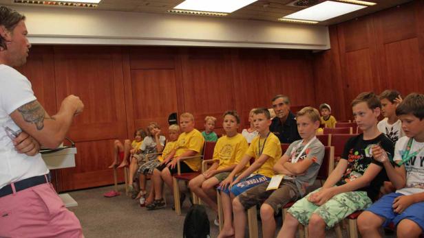 Ein Mann spricht vor einer Gruppe von Kindern in einem Raum mit Holzvertäfelung.