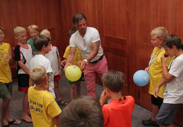 Ein Mann mit Tätowierungen erklärt einer Gruppe von Kindern etwas mit Ballons.