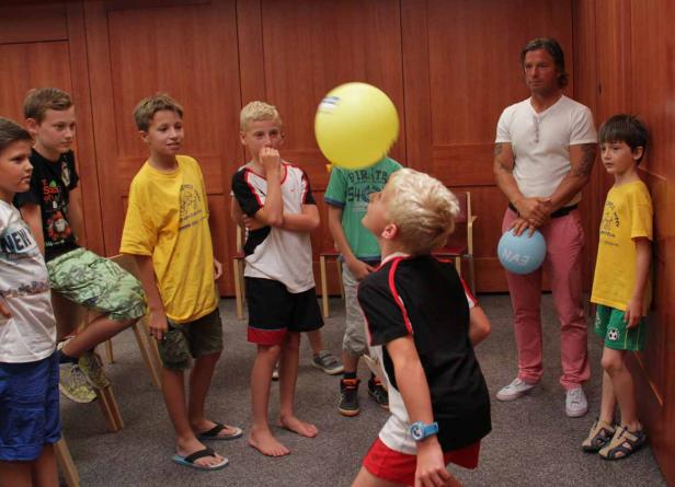 Eine Gruppe von Kindern spielt mit einem gelben Ballon in einem Raum. Ein Mann mit Tätowierungen steht daneben.