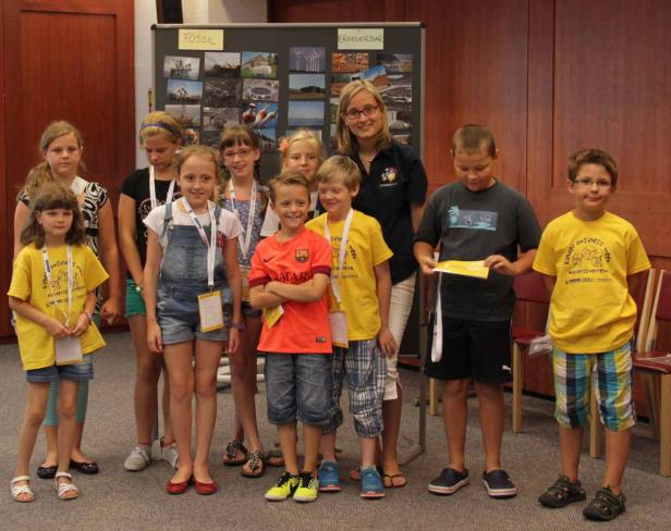 Eine Gruppe Kinder und eine Frau stehen vor einer Tafel mit Bildern zu den Themen Fossilien und erneuerbare Energien.