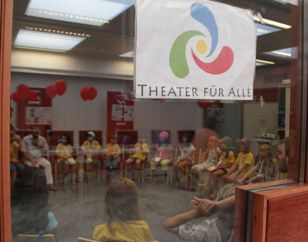 Eine Gruppe Kinder sitzt im Kreis in einem Raum, gesehen durch ein Fenster mit dem Schild „Theater für Alle“.