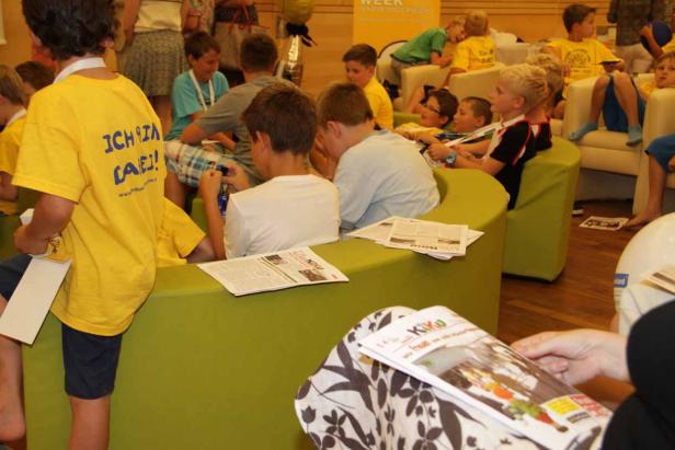 Eine Gruppe Kinder sitzt auf grünen Sitzgelegenheiten und liest Zeitungen.