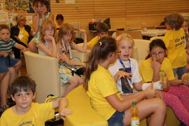 Eine Gruppe Kinder sitzt und steht in einem Raum, einige tragen gelbe T-Shirts.