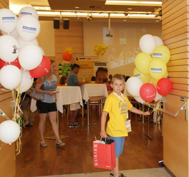 Ein Junge mit Luftballons und einer Tasche von „Kurier“ betritt einen Veranstaltungsraum.