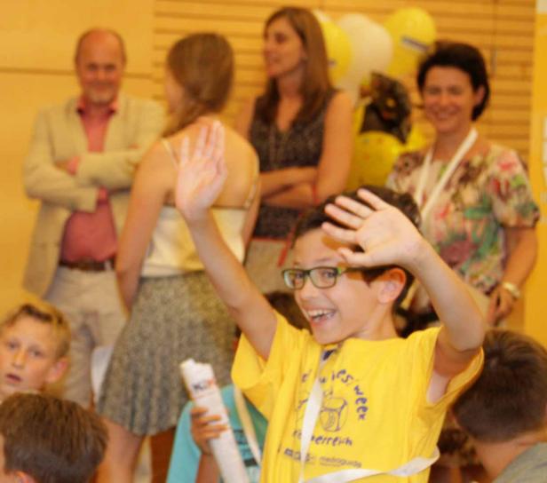 Ein lächelnder Junge mit Brille winkt in die Kamera, umgeben von anderen Personen.
