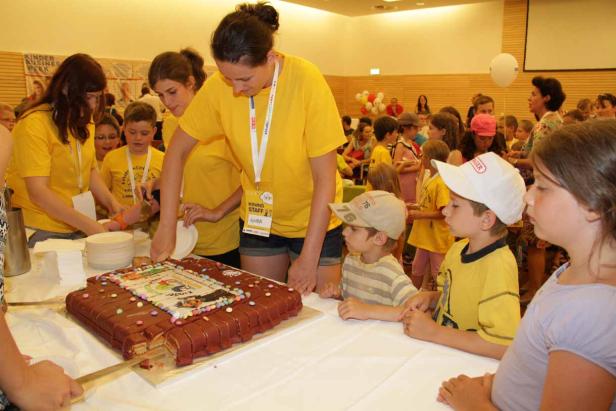 Kinder stehen um einen Tisch, an dem ein großer, rechteckiger Kuchen angeschnitten wird.