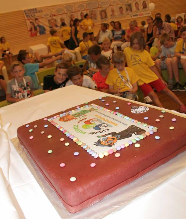 Eine große, rechteckige Torte mit dem Logo der „Kinder Business Week“ und vielen Kindern im Hintergrund.