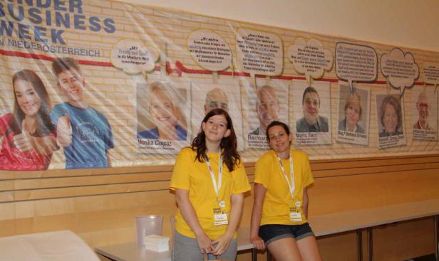 Zwei Frauen in gelben T-Shirts stehen vor einem Banner der „Kinder Business Week“.