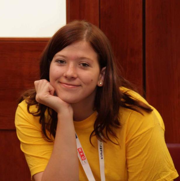 Eine junge Frau mit braunen Haaren lächelt und stützt ihr Kinn auf ihre Hand.