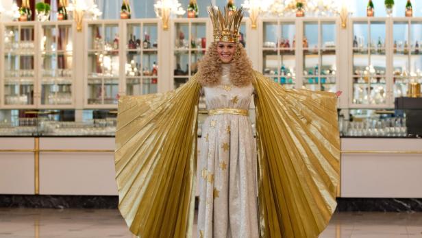 Eine junge Frau in einem goldenen Kleid und einer Krone steht vor einem Regal mit Spirituosen.