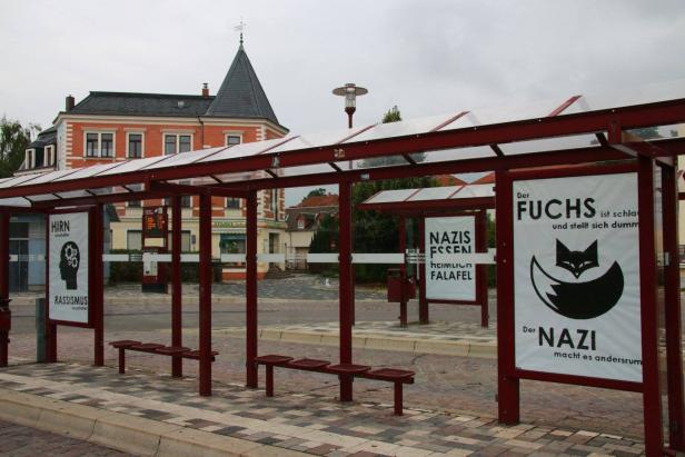 Eine Bushaltestelle mit satirischen Plakaten gegen Rassismus und Nazis, darunter ein Fuchs-Motiv.