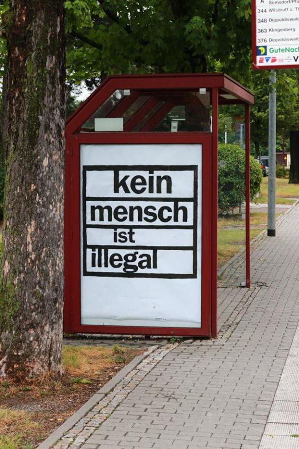 Ein rotes Wartehäuschen zeigt ein Plakat mit der Aufschrift „Kein Mensch ist illegal“.
